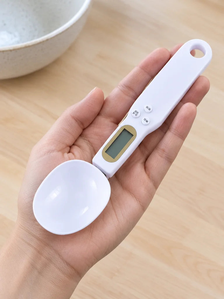 A close kitchen photograph of the complete white Doseo digital weighing spoon resting across an open