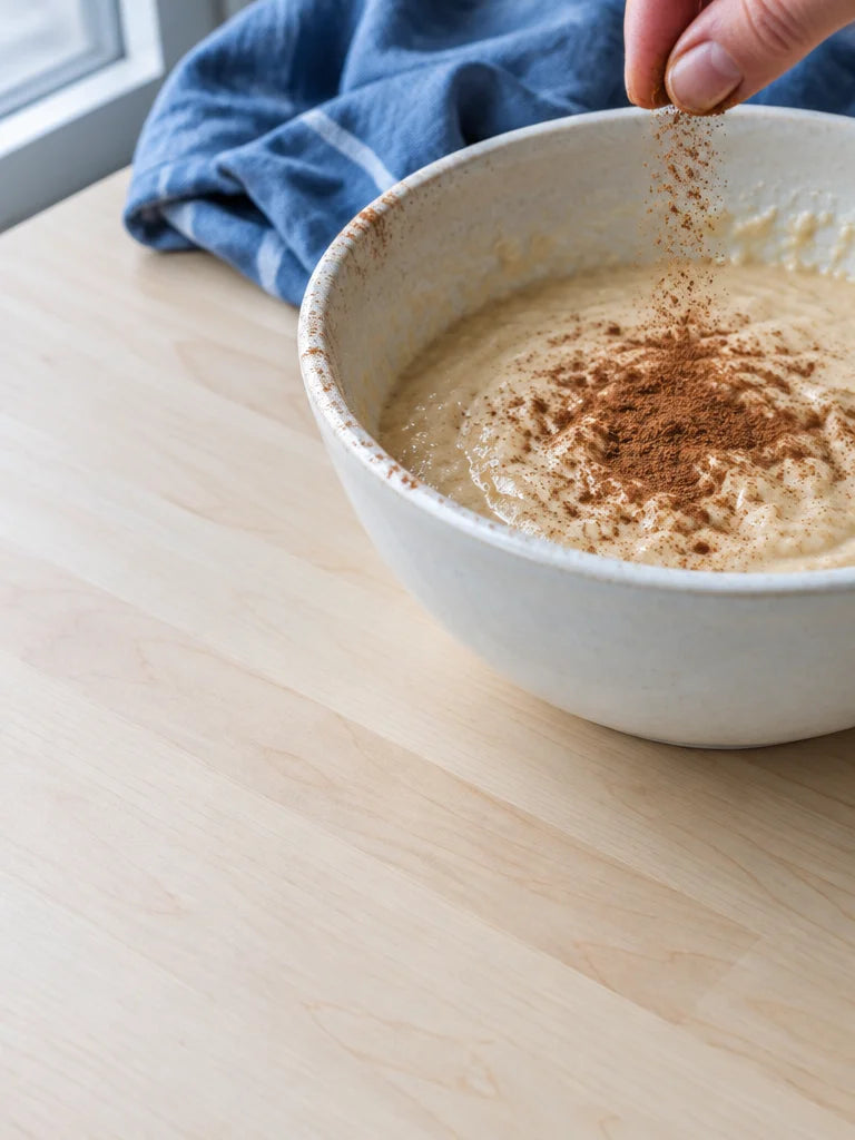 A close kitchen photograph of a cinnamon pinch falling unevenly from fingertips into a pale ceramic